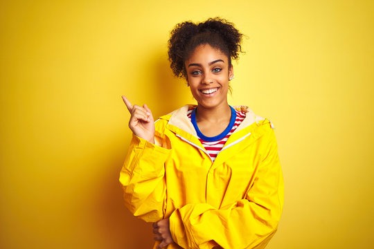 Young african american woman wearing rain coat over isolated yellow background with a big smile on face, pointing with hand and finger to the side looking at the camera.