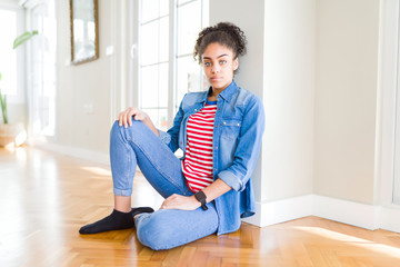 Beautiful young african american woman with afro hair sitting on the floor with serious expression...