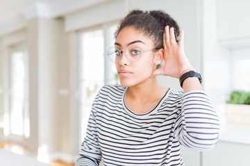 Beautiful young african american woman with afro hair wearing glasses smiling with hand over ear listening an hearing to rumor or gossip. Deafness concept.