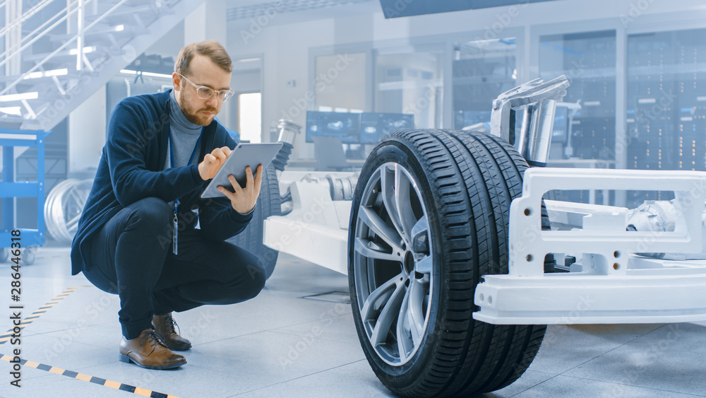 Poster Engineer with Glasses and Beard Works on a Tablet Computer Next to an Electric Car Chassis Prototype with Wheels, Batteries and Engine in a High Tech Development Laboratory.