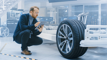 Engineer with Glasses and Beard Works on a Tablet Computer Next to an Electric Car Chassis...