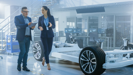 Female and Male Engineer Walk in a High Tech Development Facility Holding a Tablet Computer and Pass an Electric Car Chassis Prototype with Batteries and Wheels.
