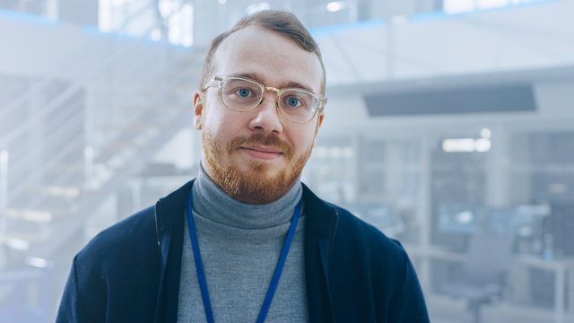 Handsome Ginger Creative Design Engineer With Beard And Glasses Poses For Camera Next To An Electric Car Chassis Prototype. In High Tech Laboratory Facility With Vehicle Frame.