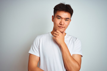 Fototapeta premium Young asian chinese man wearing t-shirt standing over isolated white background looking confident at the camera smiling with crossed arms and hand raised on chin. Thinking positive.