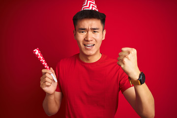 Asian chinese man on birthday celebration wearing funny hat over isolated red background annoyed and frustrated shouting with anger, crazy and yelling with raised hand, anger concept