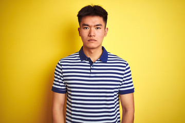 Young asian chinese man wearing striped polo standing over isolated yellow background Relaxed with serious expression on face. Simple and natural looking at the camera.