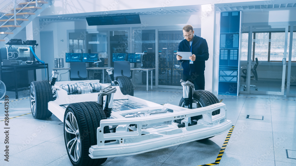 Leinwandbilder Engineer with Glasses and Beard Scans Electric Car Chassis Prototype with Wheels, Batteries and Engine with an Augmented Reality Software on a Tablet Computer in a High Tech Development Laboratory.