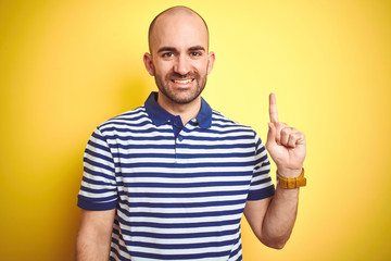 Young bald man with beard wearing casual striped blue t-shirt over yellow isolated background showing and pointing up with finger number one while smiling confident and happy.