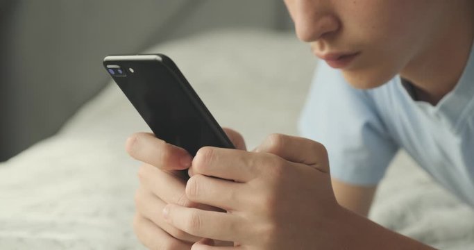 Teenager typing text message by smart phone, at home. Handsome boy spending time in a social network using mobile phone lying on sofa. Caucasian teenage guy talking on cell phone. Lifestyle concept.