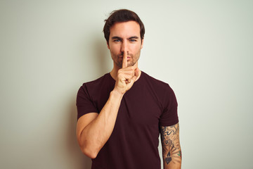 Young handsome man with tattoo wearing purple casual t-shirt over isolated white background asking to be quiet with finger on lips. Silence and secret concept.