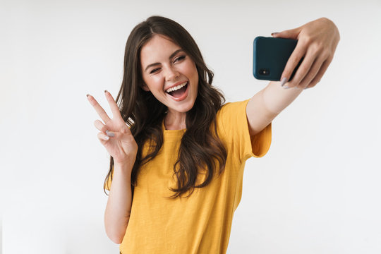 Image of beautiful brunette woman laughing and showing peace sign while taking selfie photo on cellphone