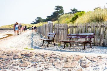 People walking in family or between friends on the GR34 long-distance footpath along the beach in Penvenan, Brittany, on a sunny summer morning with two empty wooden benches in the foreground.