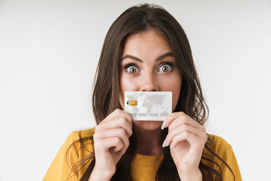 Image of surprised brunette woman wearing casual clothes holding credit card at her mouth