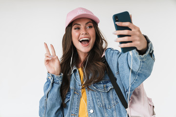 Photo of excited student girl wearing cap and earphones smiling while showing peace sign at smartphone