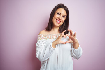 Young beautiful brunette woman over pink isolated background smiling in love showing heart symbol and shape with hands. Romantic concept.