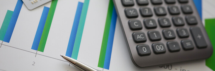 Fototapeta premium Silver pen gray calculator and credit plastic card lie on table aganist business chart background.