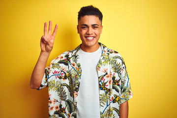 Young brazilian man on vacation wearing summer floral shirt over isolated yellow background showing and pointing up with fingers number three while smiling confident and happy.