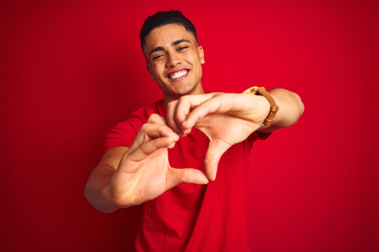 Young brazilian man wearing t-shirt standing over isolated red background smiling in love doing heart symbol shape with hands. Romantic concept.