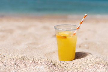 Glass of refreshing lemonade with a tube on a sand beach with blue ocean background. Trip and summer relax concept. Copy space.