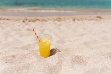 Glass of refreshing lemonade with a tube on a sand beach with blue ocean background. Trip and summer relax concept. Copy space.