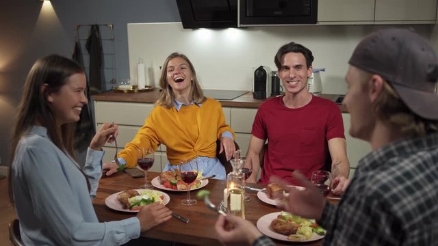 Group of four young friends dining and having fun together in domestic kitchen. Friends sitting at table talking and laughing over dinner