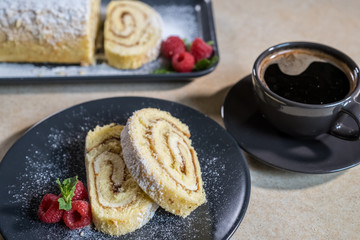Swiss Roll with jam, decorated with raspberries and mint on a plate and black coffee. Selective focus.