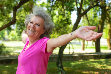 Positive carefree old lady enjoying morning exercise outdoors. Senior grey haired woman in casual standing in park, spreading hands and smiling. Active senior woman concept