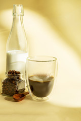 Morning coffee concept. Stylish poster for coffee. Bottle with milk and double-walled glass with poured coffee and grain on yellow background with shadows. Copy space. Flat lay food