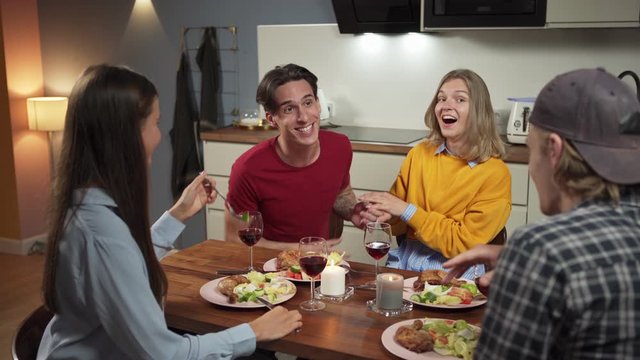 Group of four happy friends, two couples, dining in domestic kitchen. Young people talking and laughing together over dinner