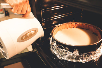 Cheesecake in the oven. Springform pan in foil. Woman is pouring water from kettle. Baking process....