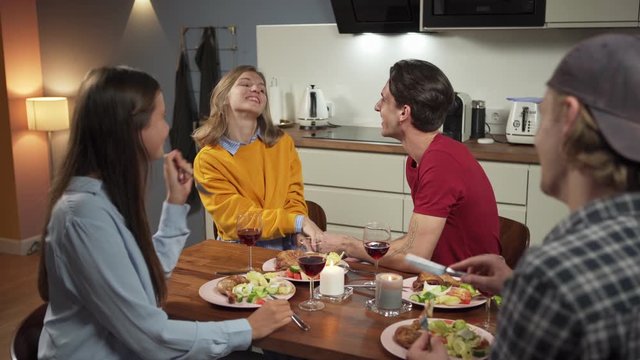 Group of four cheerful friends, two couples, having dinner in domestic kitchen. Young people eating, talking and laughing together