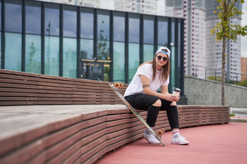 Fototapeta premium Image of brunette sportswoman in sunglasses with glass in her hands sitting on wooden bench near skateboard on summer day