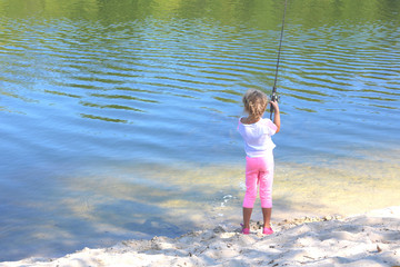 Little child fishing in summer on river on sandy shore against background of blue water and displaying green trees in river