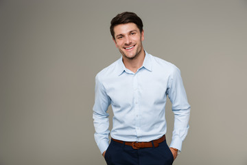 Image of happy brunette man wearing formal clothes smiling at camera with hands in pockets - obrazy, fototapety, plakaty