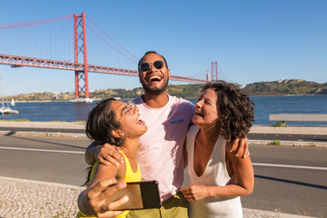 Happy close friends enjoying meeting and taking group selfie outdoors. Man and women standing on promenade, using smartphone, laughing out loud and hugging. Fun with friends concept