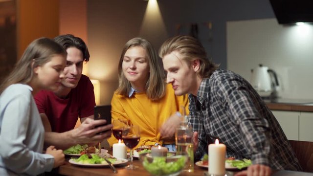 Zoom out shot of four young friends having dinner in domestic kitchen. Man showing photos or videos on cell phone, friends watching and laughing