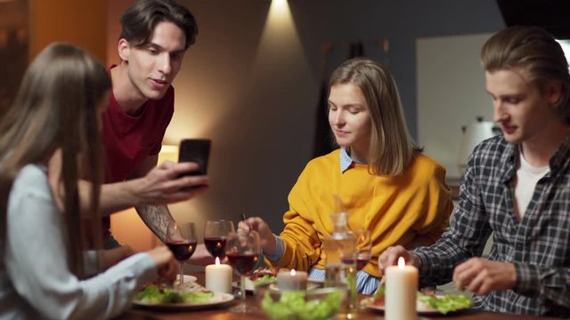 Zoom out shot of four young friends having dinner together. Man showing funny pictures or videos on cell phone, friends watching and having fun