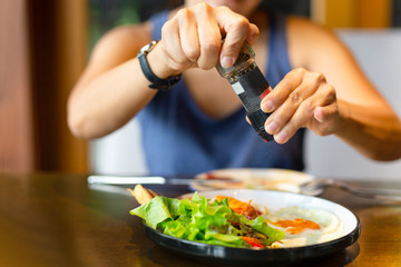 woman grinding pepper over fried eggs at breakfast with glass of water on table.