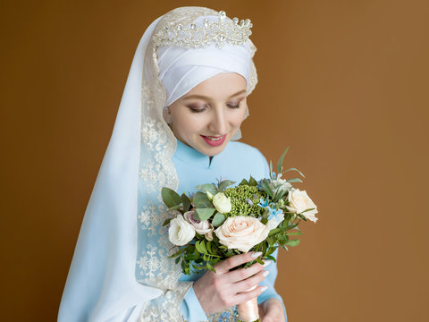 Portrait of beautiful muslim bride with bouquets hands modestly looks down on brown background in studio