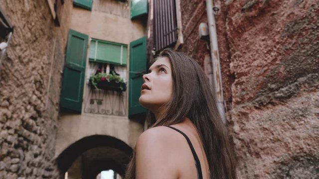 Beautiful girl with long brown hair and black top looking up among old walls of colorful houses exploring old town torri del benaco on lake garda, veneto italy august summer 2019