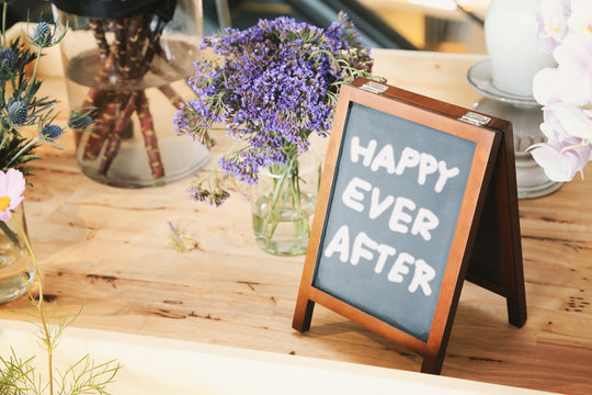 The words "HAPPY EVER AFTER" on small blackboard with easel among flowers vase on wooden table, toned image.