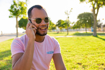 Happy excited Latin guy in sunglasses speaking on mobile phone in park. Young man in casual sitting on grass and calling on cellphone. Phone talk outdoors concept