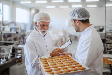 Young Caucasian employee holding tray with fresh cookies while supervisor evaluating quality and holding tablet. Both are dressed in sterile white uniforms and having hairnets. Food plant interior.