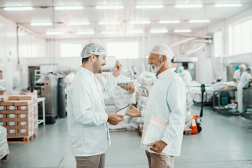 Two supervisors talking about quality of food. Younger one holding tablet while older one holding folder with charts. Both are dressed in sterile uniforms and having hairnets. Food plant interior.