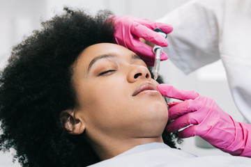 Attractive young African woman is getting a rejuvenating facial injections. She is sitting calmly...