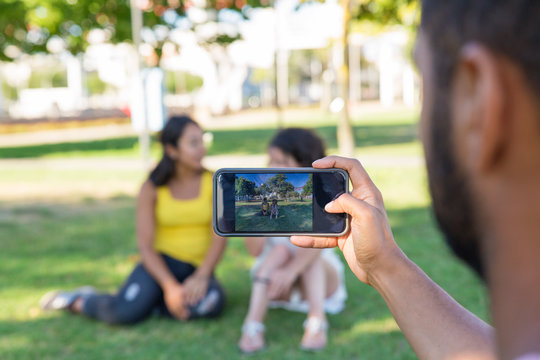 Man taking picture of two female friends on phone. Women sitting on grass in park and chatting. Photo outdoors concept