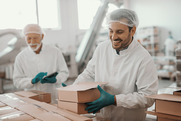 Smiling hardworking dedicated Caucasian employee arranging boxes while his superior using tablet and checking on him. Both are dressed in white sterile uniforms. Food plant interior.