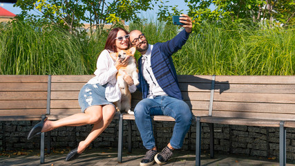 The happy couple are sitting on the bench in the park with little dog and do selfie portrait. Cute corgi Pembroke puppy on its owners hands
