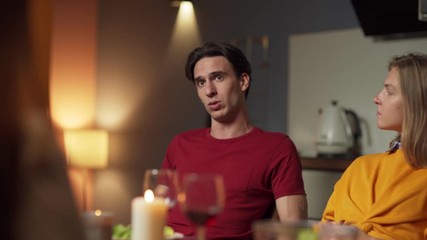 Tracking shot of group of four young people sitting at table and having dinner together. Selective focus on man talking to friends