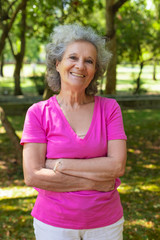 Happy joyful old lady posing outdoors. Senior grey haired woman in casual standing in park with arms folded, looking at camera and smiling. Outdoor female portrait concept